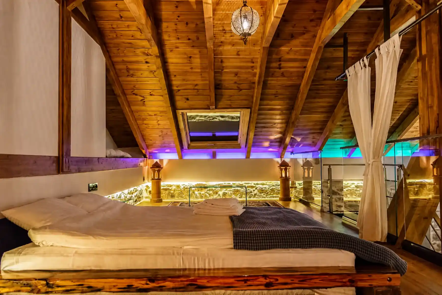 Mezzanine bedroom under a beamed timber roof, with a double bed, glass balustrade and a hanging lantern